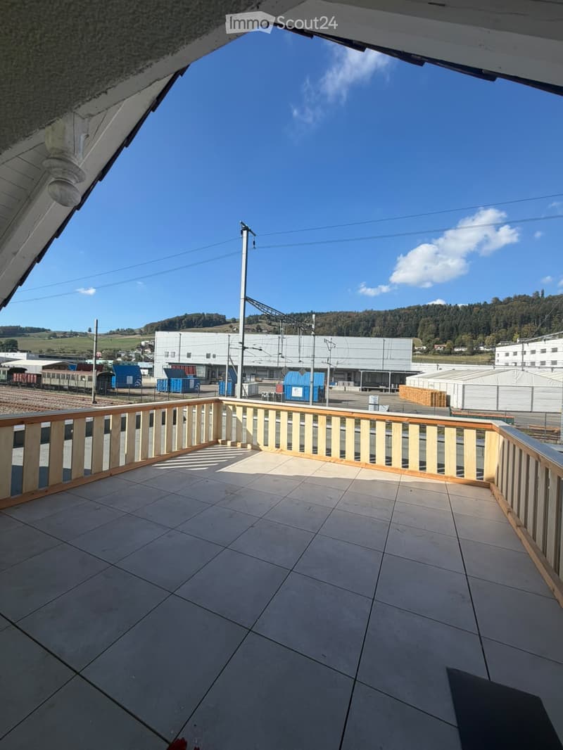 Balcony with tiled floor and wooden railing, overlooking an industrial area with buildings, train tracks, and distant hills.