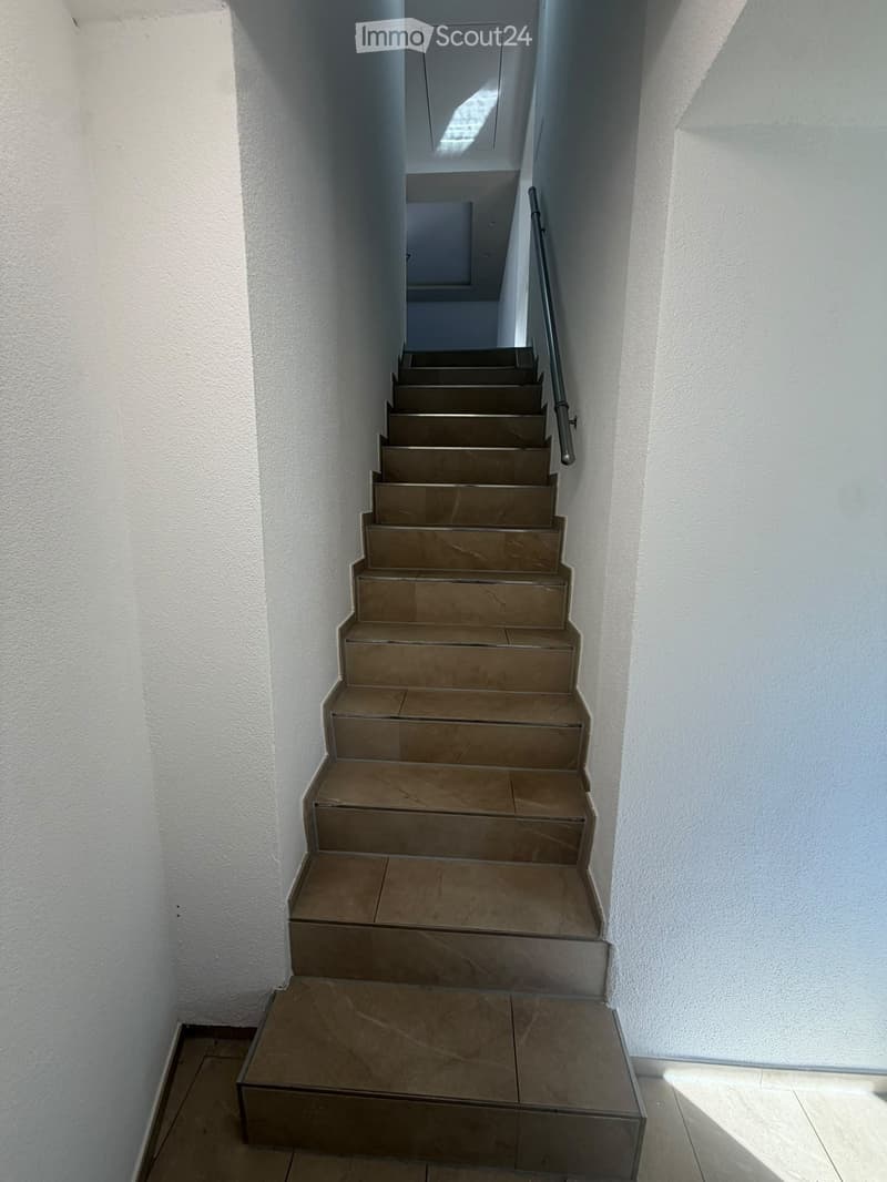 Staircase with beige tiled steps and a metal handrail, white walls, and a small skylight at the top.