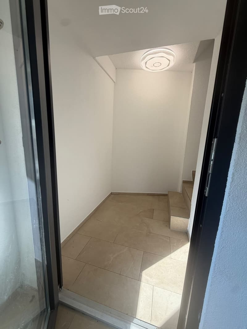 Empty hallway with tiled floor, white walls, and a ceiling light fixture. A small staircase leads upwards.