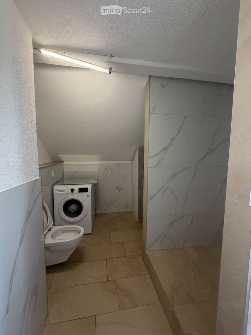 Modern bathroom with beige tiled floor, white toilet, and a private washing machine. Marble-style wall tiles and sleek cabinetry.