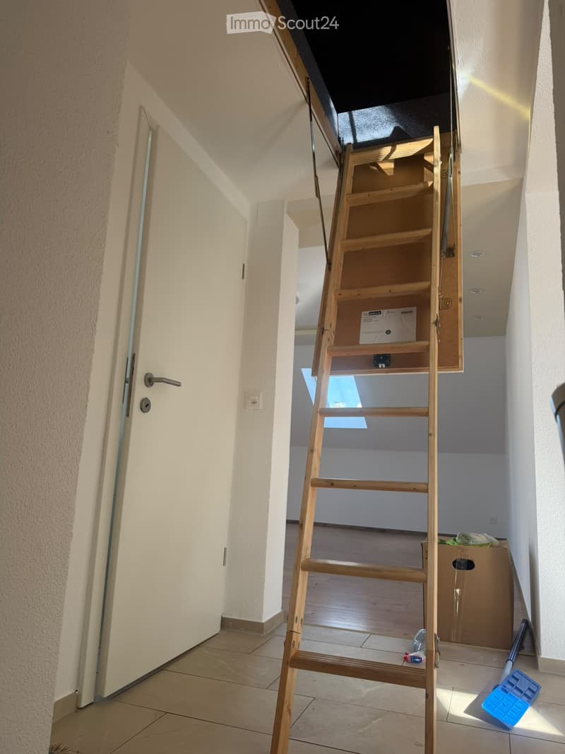 A narrow hallway with a wooden staircase leading upwards, tiled floor, white walls, and a partially open door on the left.