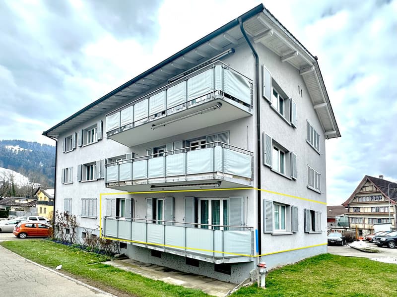 A three-story residential building with multiple balconies, white exterior, and multiple windows. The ground floor has a covered balcony area.