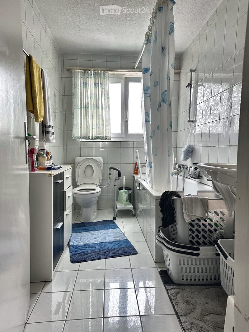 Bathroom with tiled walls and floor, toilet, bathtub, sink, shower curtain, towels, laundry basket, and a blue bath mat.