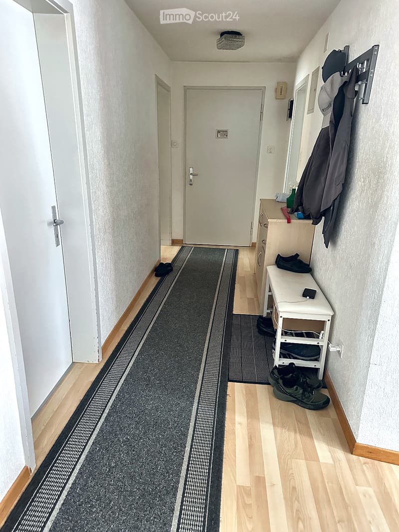 A narrow hallway with wooden flooring, a gray runner rug, and a small white shelf holding shoes and a phone. There are two doors at the end of the hallway.