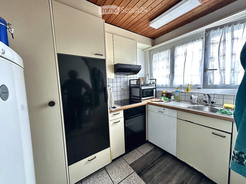 A compact kitchen with white cabinets, a refrigerator, oven, microwave, dishwasher, and a sink with a window above it. The space features a wooden ceiling and tiled walls.