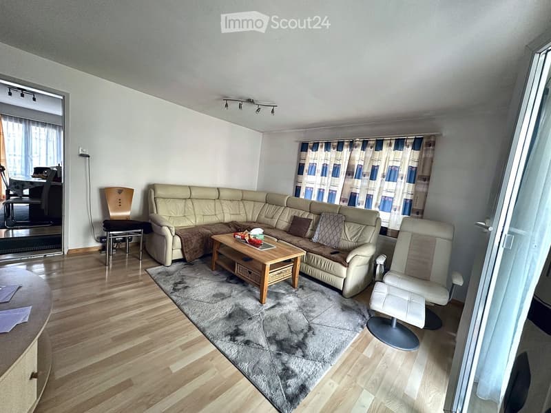 Modern living room with a beige sectional sofa, wooden coffee table, grey rug, and large windows with curtains. The room features hardwood flooring and a dining area visible in the background.