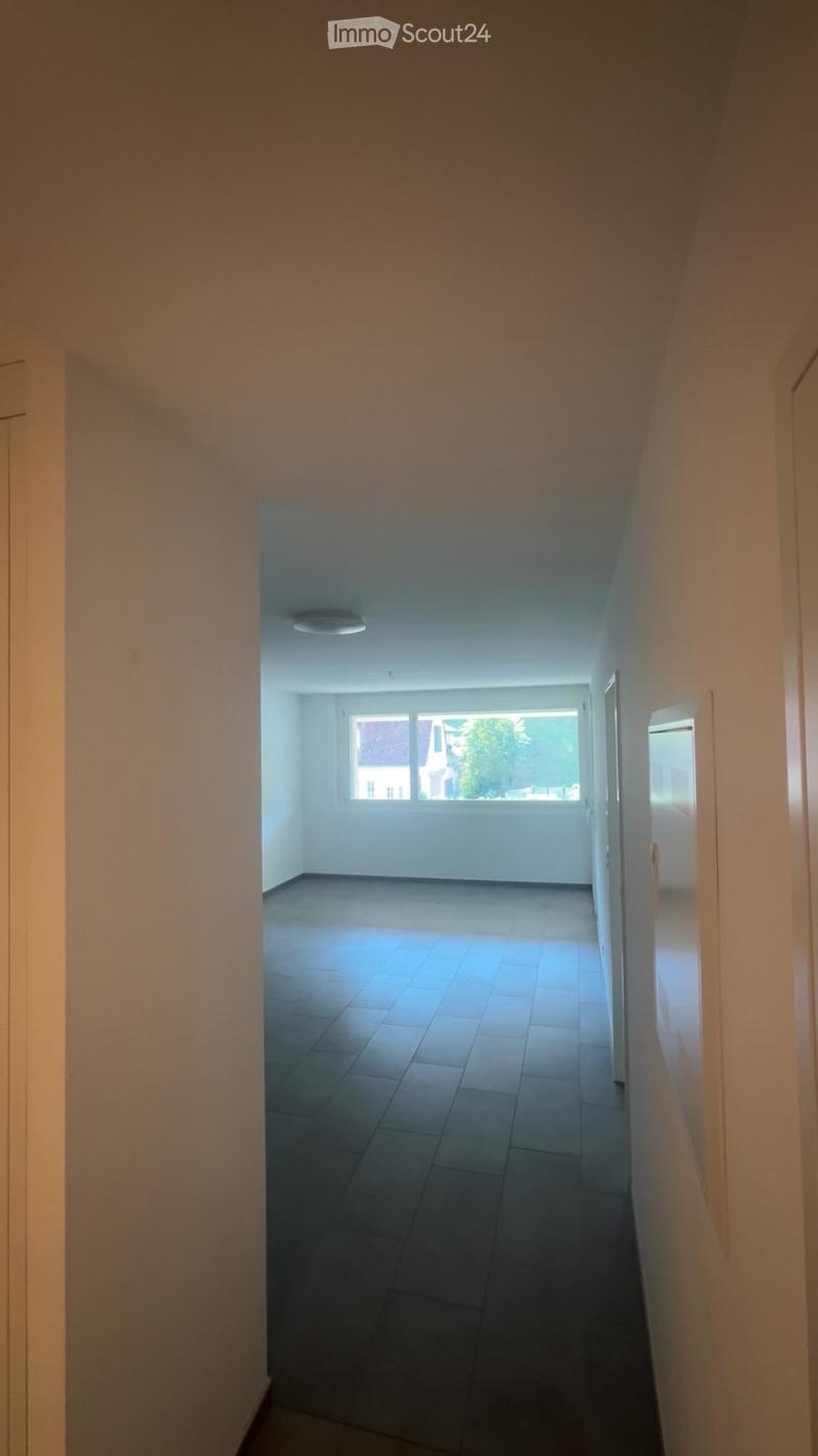 Empty hallway with tiled floor, white walls, and a window at the end showing a view of a building and greenery outside.