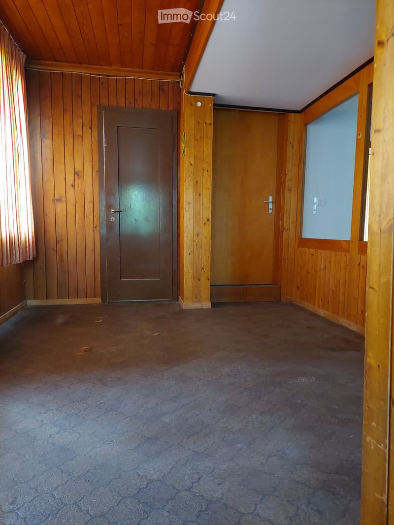 Empty room with wooden walls, wooden ceiling, and concrete floor. Features two doors and a window with a curtain.