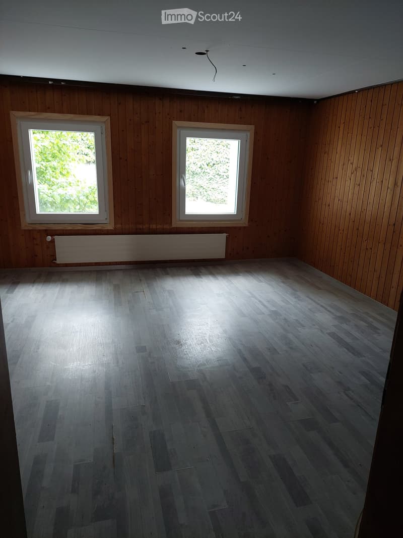 Empty room with wooden walls and floor, two windows, and a radiator. No furniture or equipment present.