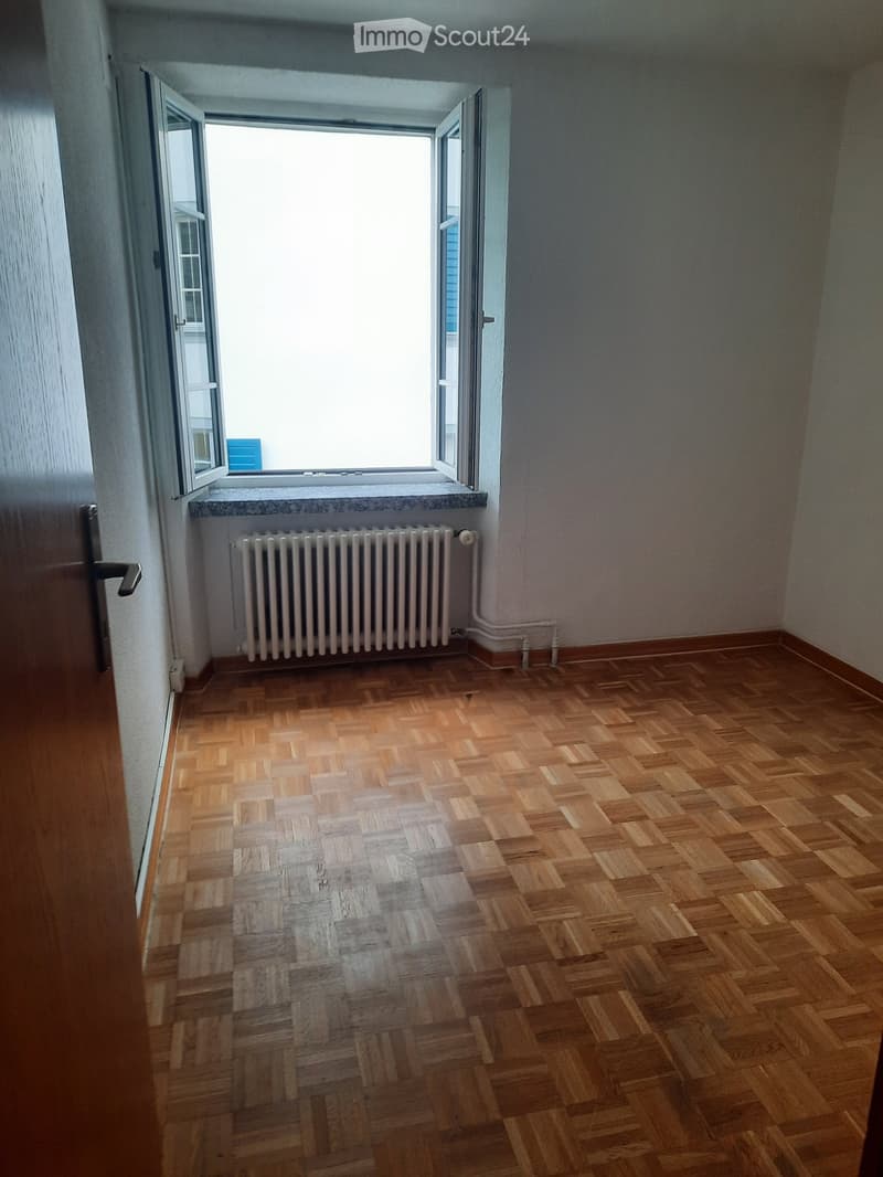 Empty room with wooden parquet flooring, white walls, a radiator, and a large window with open shutters.