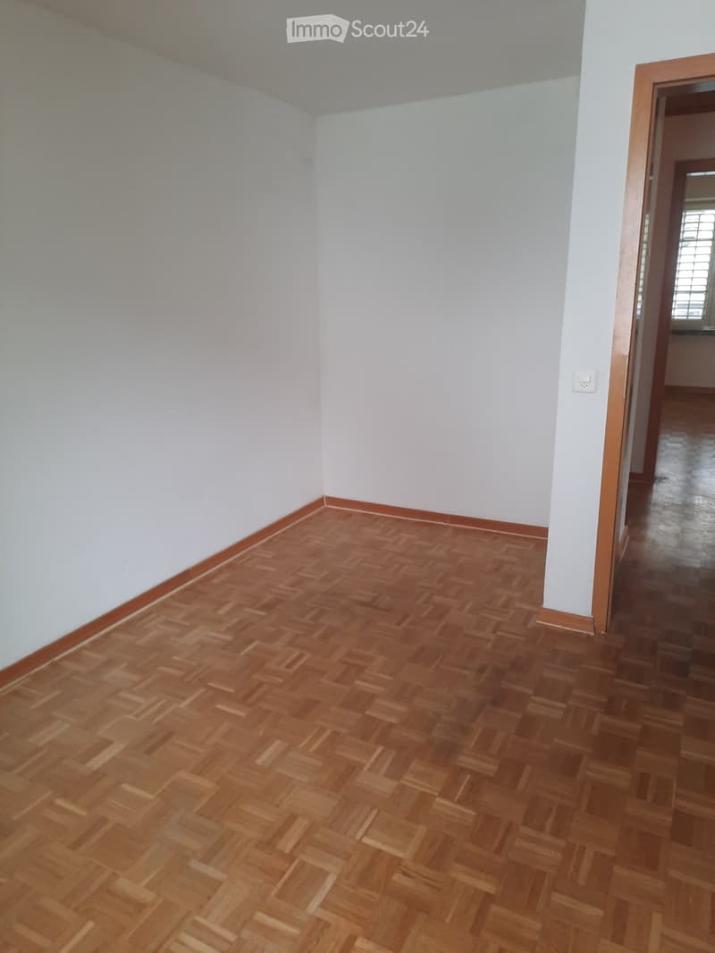 Empty room with wooden parquet flooring and white walls, featuring a doorway leading to another space.