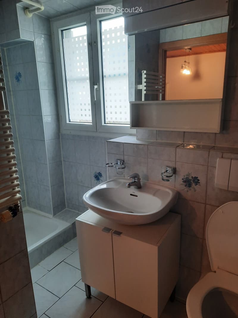 Modern bathroom with a white sink, faucet, mirror, cabinet, and toilet. The room features tiled walls and a large window with a view of a building.