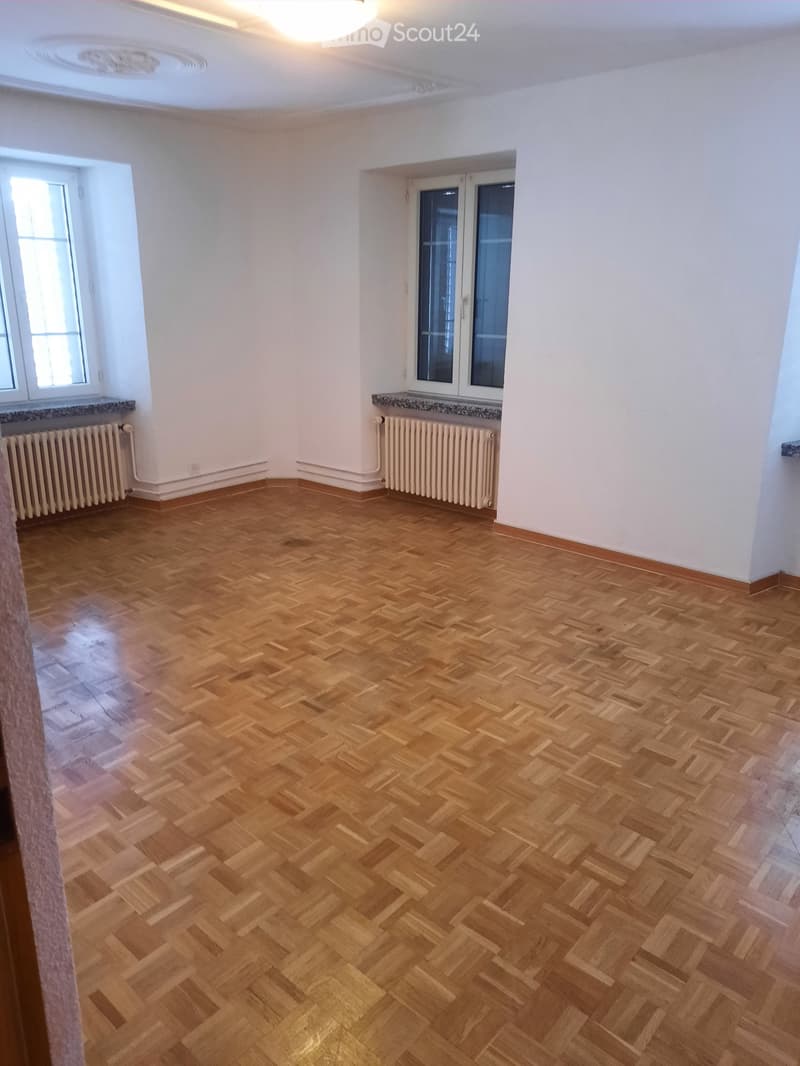 Empty room with wooden herringbone flooring, white walls, and large windows with radiators beneath them.