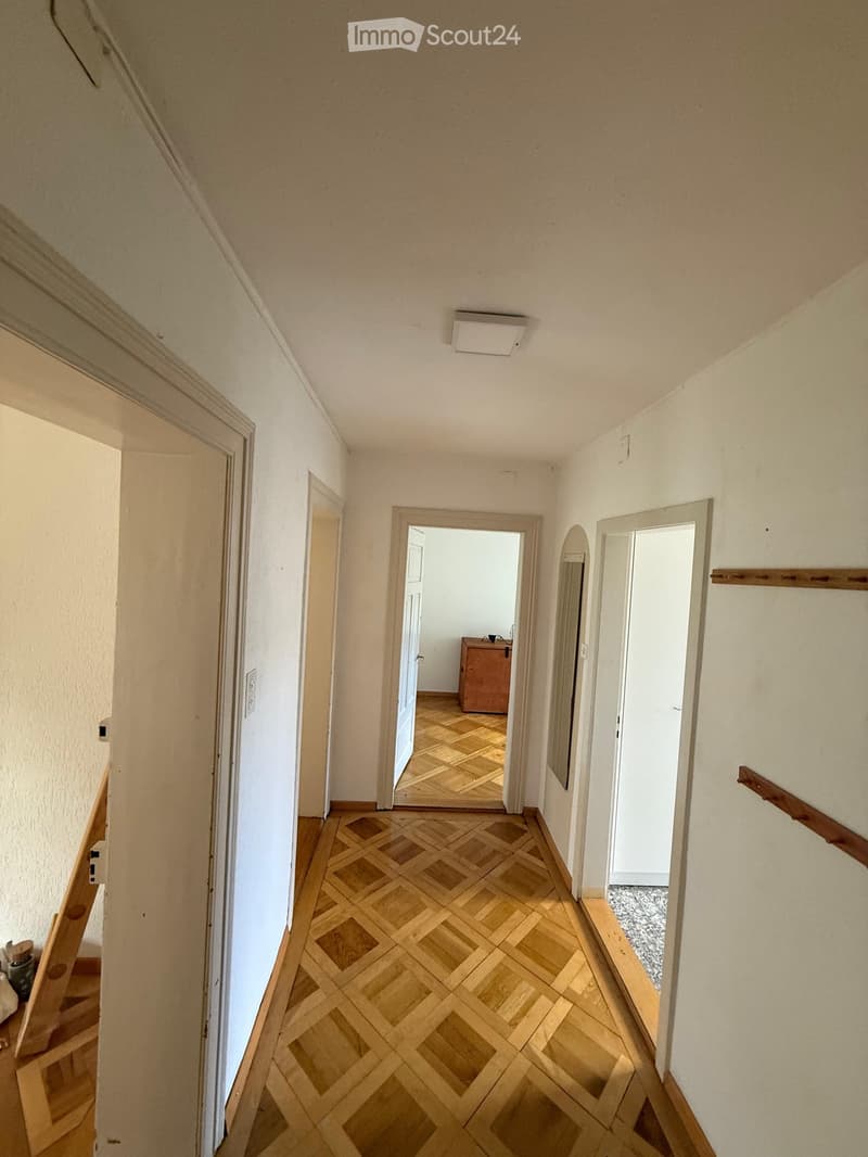 A narrow hallway with wooden parquet flooring, white walls, and several doors leading to different rooms. The hallway features a staircase on the left and a small cabinet visible through an open door at the end.