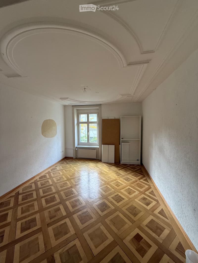Empty room with wooden parquet flooring, white walls, and a large window with a radiator below it. The ceiling features ornate molding and a decorative arched design.