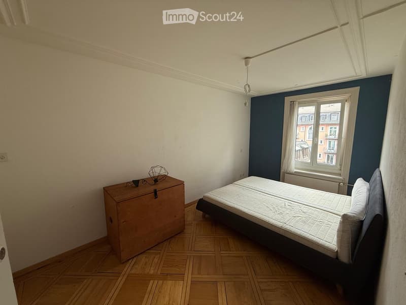 A small, furnished bedroom with a single bed, a wooden nightstand, and a lamp. The room features wooden flooring and a window with white curtains, offering a view of the city.