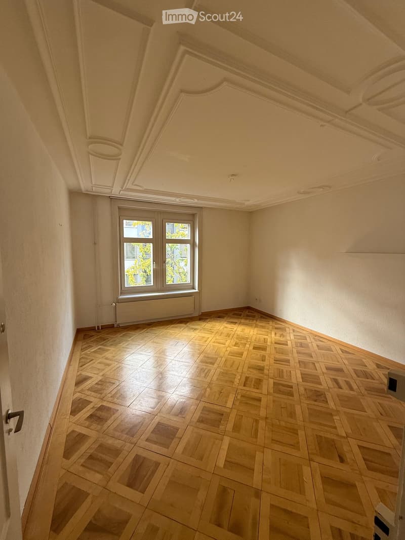 Empty room with wooden parquet flooring, white walls, and a large window with a radiator below it. The ceiling features ornate molding.