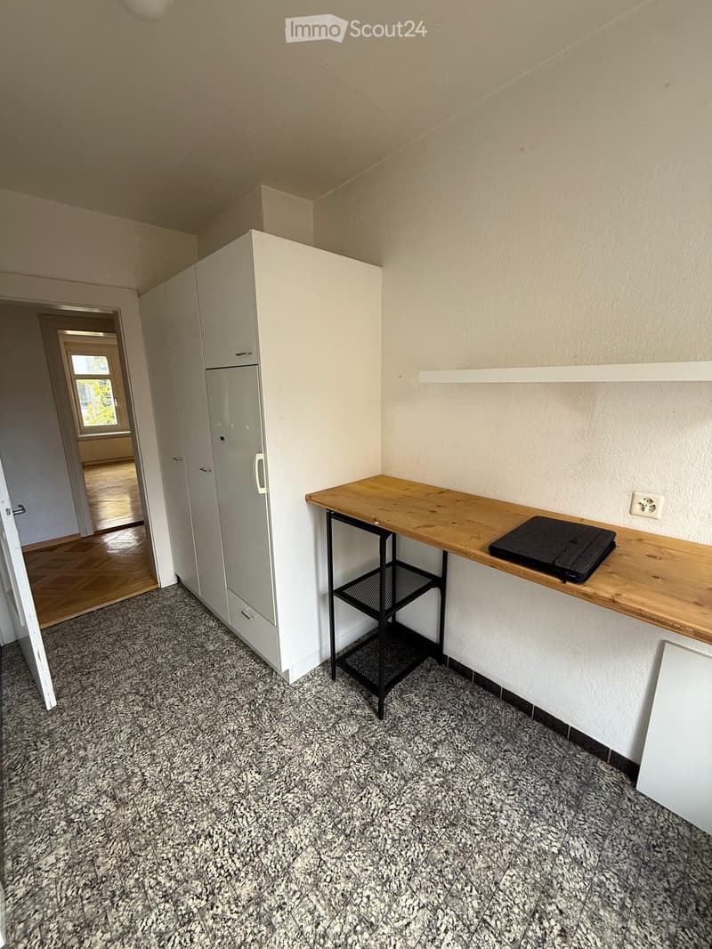 A small, modern room with a wooden table, a laptop, and a white cabinet. The floor is covered with a speckled gray tile, and the walls are painted a light beige.