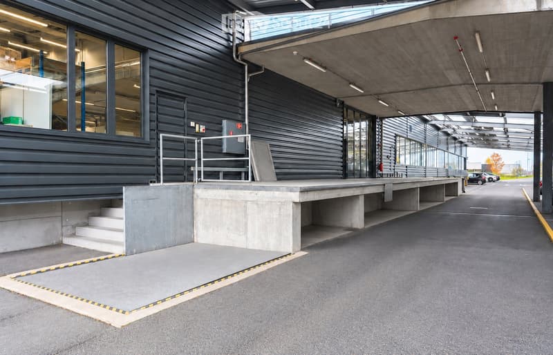 Modern building entrance with a raised ground floor, concrete platform, and glass walls. The entrance features a ramp and steps leading up to the building.
