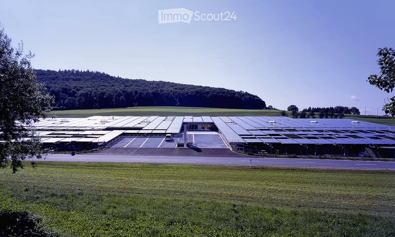 Large industrial building with solar panels on the roof, surrounded by open fields and a parking area.