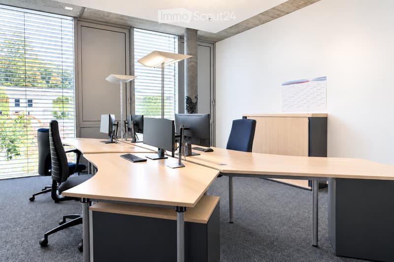 Modern office space with desks, chairs, computers, keyboards, and lamps. Large windows with blinds provide natural light.