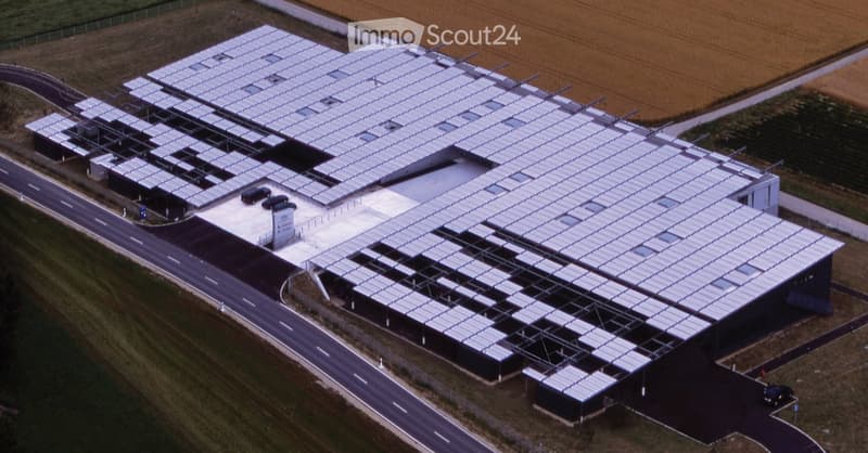 A large industrial building with a flat roof covered in solar panels, surrounded by a paved area with a few parked cars and a road nearby.