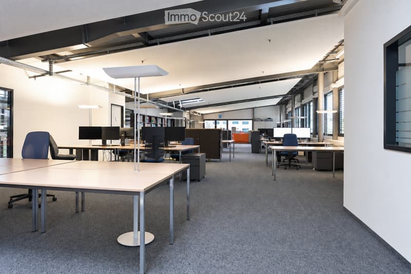 Modern office space with multiple desks, chairs, computers, and large windows allowing natural light. The area features contemporary furniture and a spacious layout.