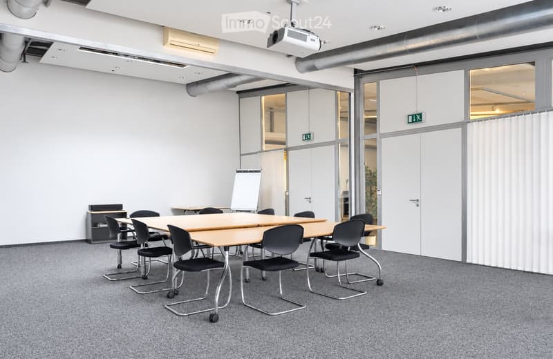 Modern meeting room with a large table, chairs, and a screen for presentations. The room features white walls, large windows, and a carpeted floor.