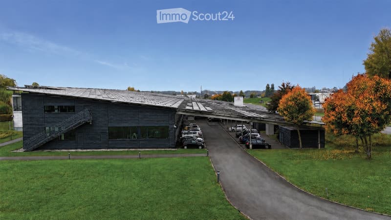 Modern building with a grey facade, large windows, and a covered parking area with several cars. The building has a sloped metal roof and is surrounded by a well-maintained lawn and trees.