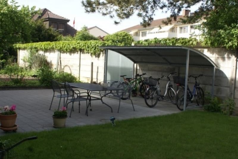 Outdoor patio area with a covered bike shelter, a table and chairs, and several bicycles. Potted plants are placed on the brick patio near the grassy area.