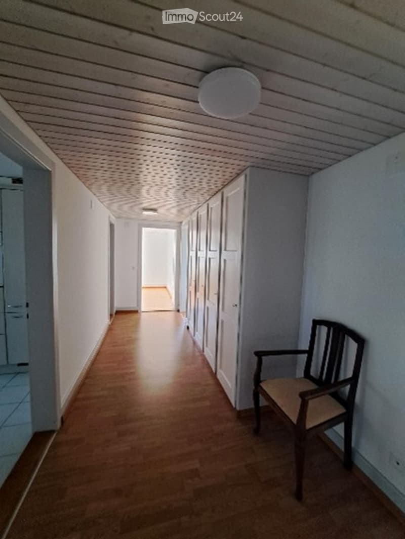 A narrow hallway with wooden flooring and white walls, featuring a single wooden chair placed against the wall.