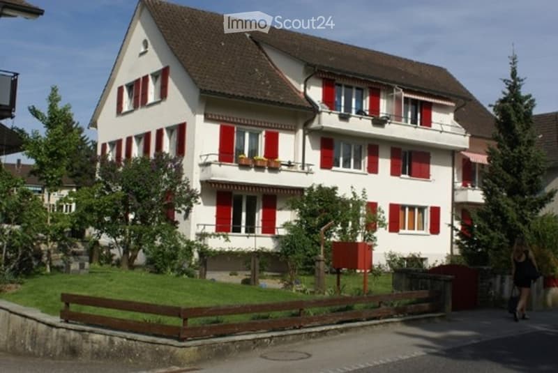 A two-story house with white walls, red shutters, and a brown roof. The house features multiple windows, balconies, and is surrounded by greenery and a wooden fence.
