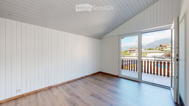 Empty room with white paneled walls, wooden flooring, and a large sliding glass door leading to a balcony with a view of mountains and a swimming pool.