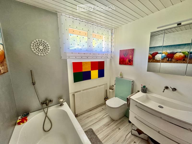 Bathroom with a bathtub, toilet, sink, mirror, and a radiator. The room features a window with a curtain, a decorative wall piece, and colorful artwork.