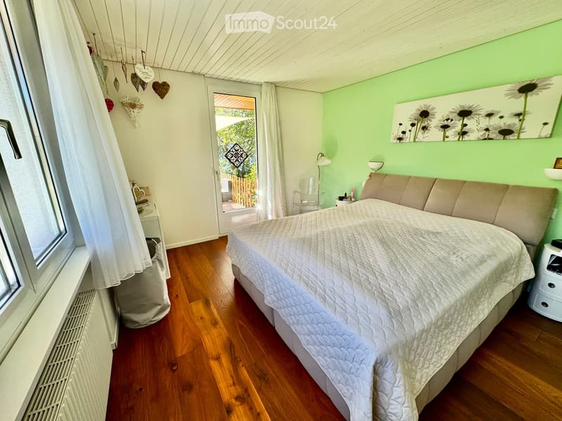 Cozy bedroom with wooden flooring, a large bed with a quilted duvet, a sofa, a nightstand with a lamp, a chair, and a large mirror on the wall. A window with a curtain and a door leading to an outdoor area are visible.