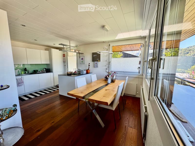 Open-plan kitchen and dining area with wooden flooring, white cabinets, a central dining table with chairs, and large windows offering a scenic view.