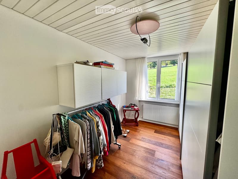 A well-lit room with wooden flooring, a wardrobe, a clothing rack, a small table with a chair, and a window with curtains.
