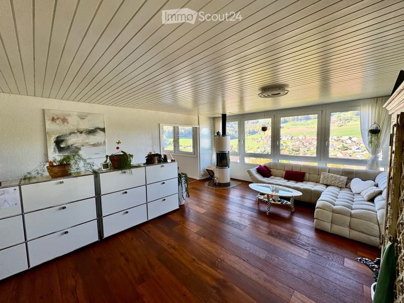 Modern living room with wooden flooring, large windows offering a scenic view, a sectional sofa, coffee table, fireplace, storage unit, and decorative plants.