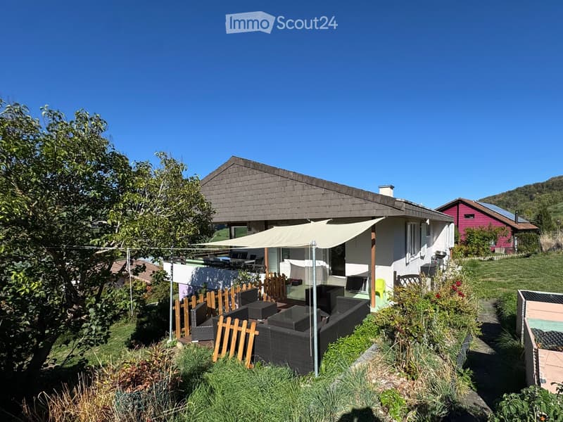 A modern house with a spacious outdoor seating area featuring a sofa, table, chairs, and a large umbrella, surrounded by greenery and a wooden fence.