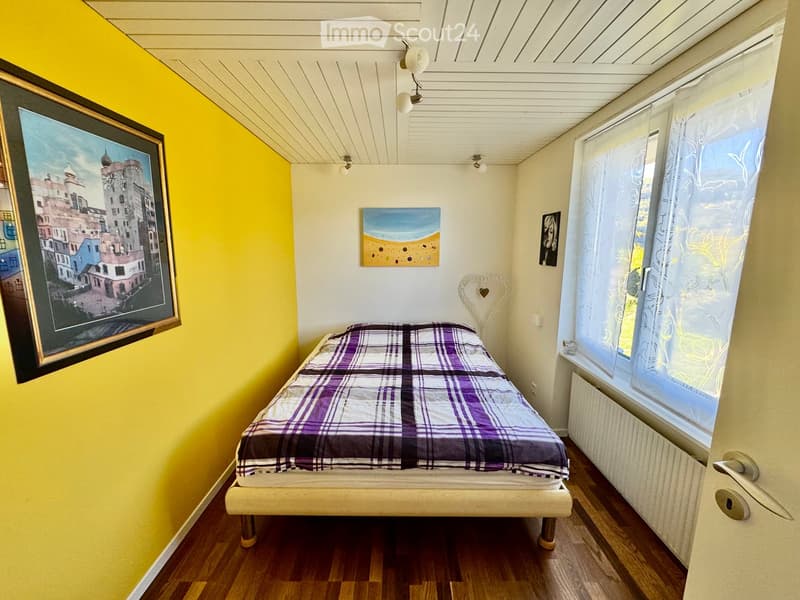Cozy bedroom with a single bed, wooden flooring, and colorful wall art. Features a large window with sheer curtains and a radiator below.
