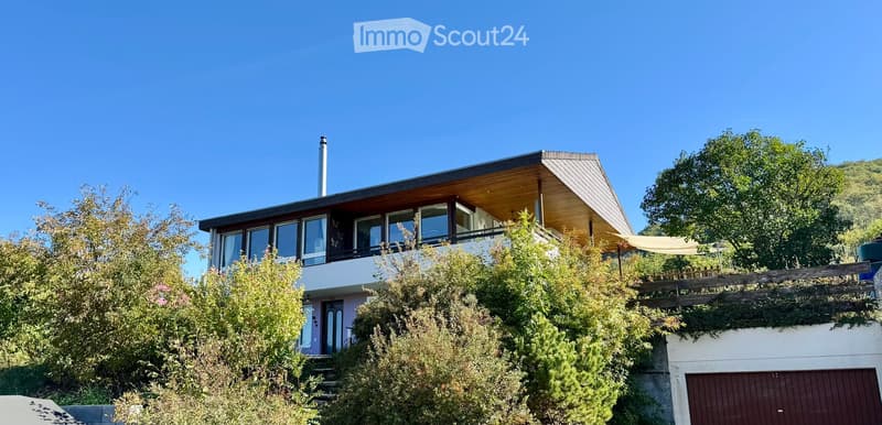 Modern two-story house with large windows, a flat roof, and a garage. The house is surrounded by greenery and has a raised ground floor.