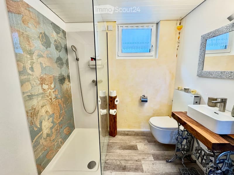 Modern bathroom with a glass-enclosed shower, wooden toilet paper holders, a white toilet, a sink with a wooden vanity, and a decorative mirror.