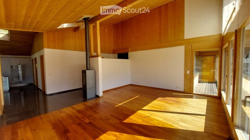 Bright, open living space with wooden floors, exposed wooden beams, and large windows. Features a modern wood-burning stove and a sliding glass door leading to a deck.