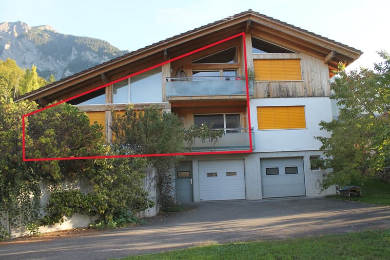 Two-story house with a wooden roof, large windows, and a balcony. Features include a garage, surrounding greenery, and a scenic mountain view in the background.