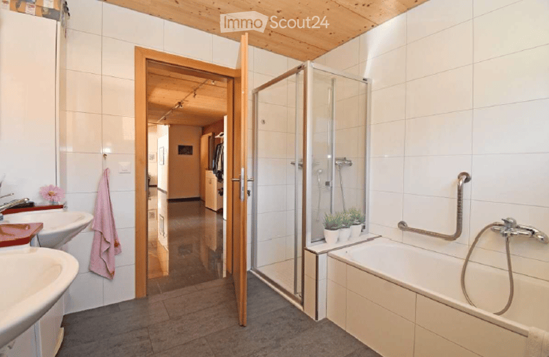 Bathroom with a bathtub, sink, and a glass-enclosed shower. A pink towel hangs on the wall, and small potted plants are placed on the edge of the bathtub. The room features white tiled walls and a wooden ceiling.