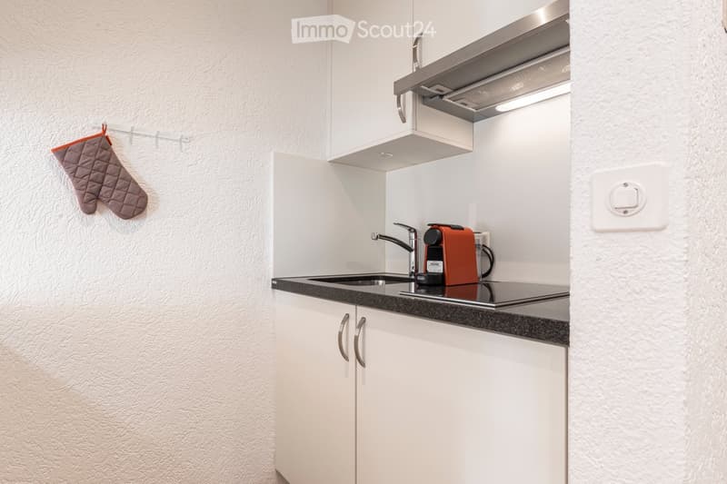 Modern kitchen with white cabinets, black countertop, stainless steel sink, coffee maker, and an oven. A gray oven mitt hangs on the wall.