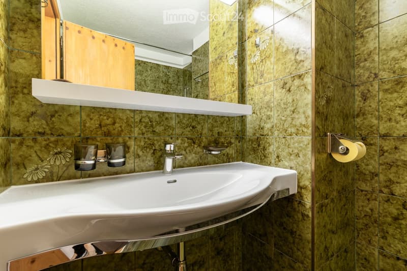 Modern bathroom with a white sink, chrome faucet, toothbrush holders, and a toilet paper holder mounted on marble-patterned walls. A wooden cabinet and a white shelf are also visible.