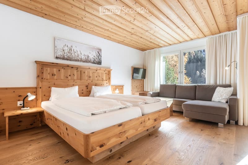 Cozy bedroom with wooden bed, nightstand, sofa, and TV, featuring large windows with a scenic view.