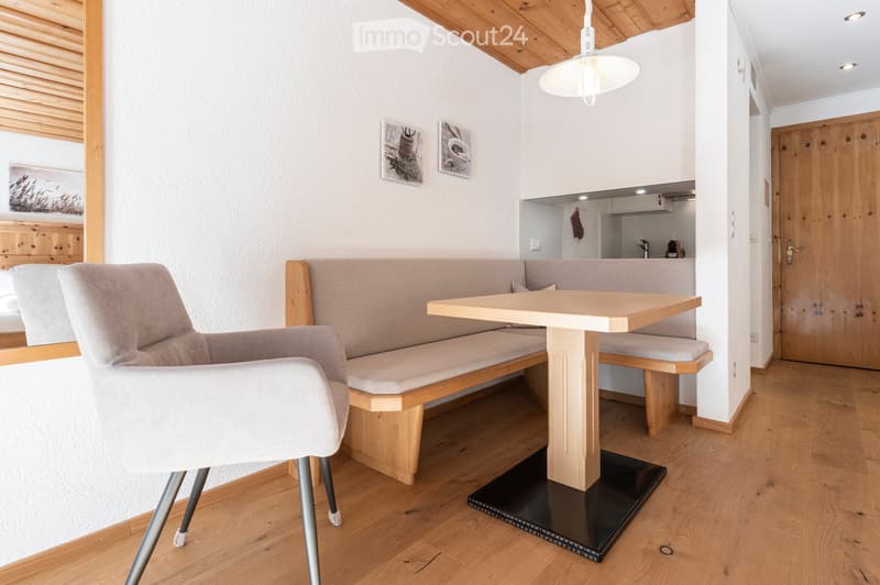 Cozy dining area with a wooden table, bench seating, and a single chair, featuring a modern pendant light and a small kitchen area in the background.