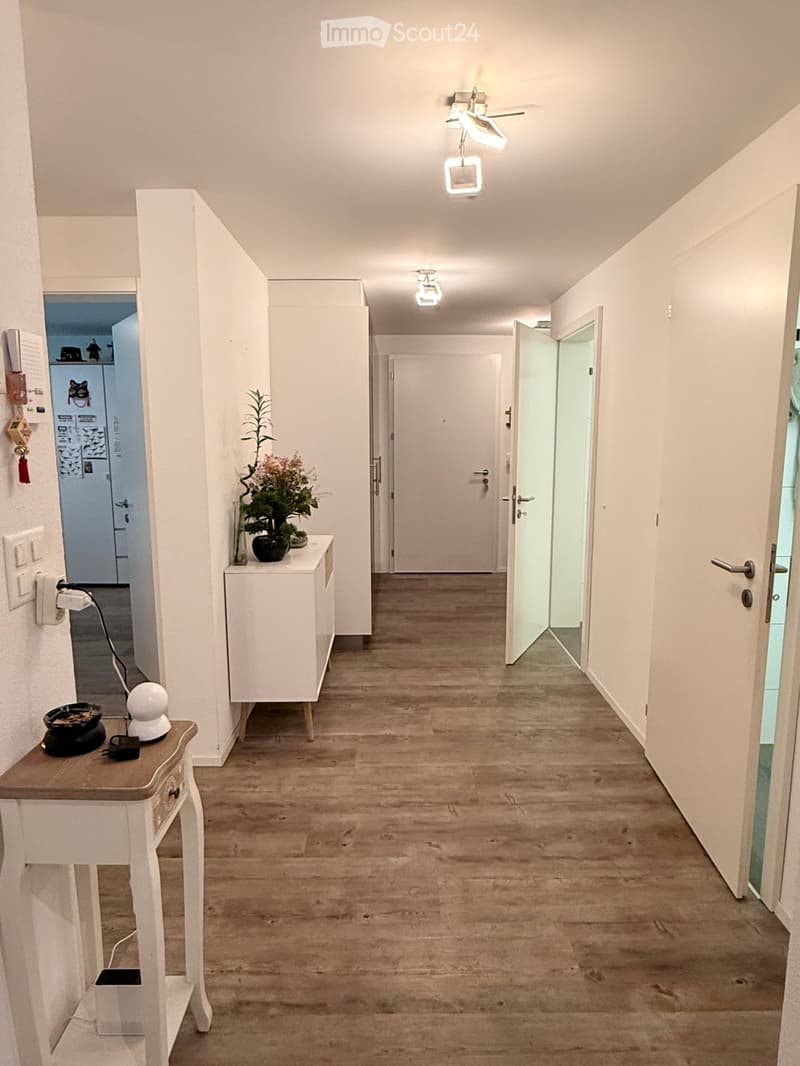 A well-lit hallway with wooden flooring, white walls, and several doors leading to different rooms. There are small tables with decorative items and a small plant on one side.